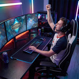 The image features a gaming chair, specifically the UVI Sport XL in white. It is designed for ergonomic comfort, suitable for long gaming sessions. In the scene, a person is celebrating a victory while seated at a gaming desk with multiple monitors, a keyboard, and a gaming mouse, surrounded by colorful LED lighting and energy drinks.