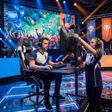 The image features a UVI gaming chair, specifically the model "UVI Sport XL" in white. It depicts a competitive gaming environment where a player is seated at a gaming desk, focused on the screen, while a teammate adjusts the monitor. The backdrop includes a lively audience and colorful stage lights, emphasizing the energetic atmosphere of the event.