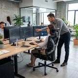 The image features an ergonomic office chair, specifically the UVI Focus Pro model. It shows a modern office environment where a woman is seated at a desk with multiple monitors, while a man is standing beside her, pointing at the screen. The workspace is well-lit and includes plants, creating a productive atmosphere.