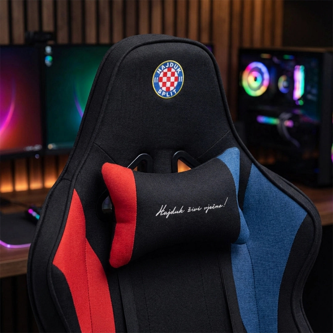 The image features a gaming chair, specifically the UVI X Hajduk model. It showcases a sleek design with a black base and colorful accents in red and blue, along with a decorative cushion that has the phrase "Hajduk živi vječno!" The chair is positioned in front of a gaming setup with monitors in the background, highlighting its