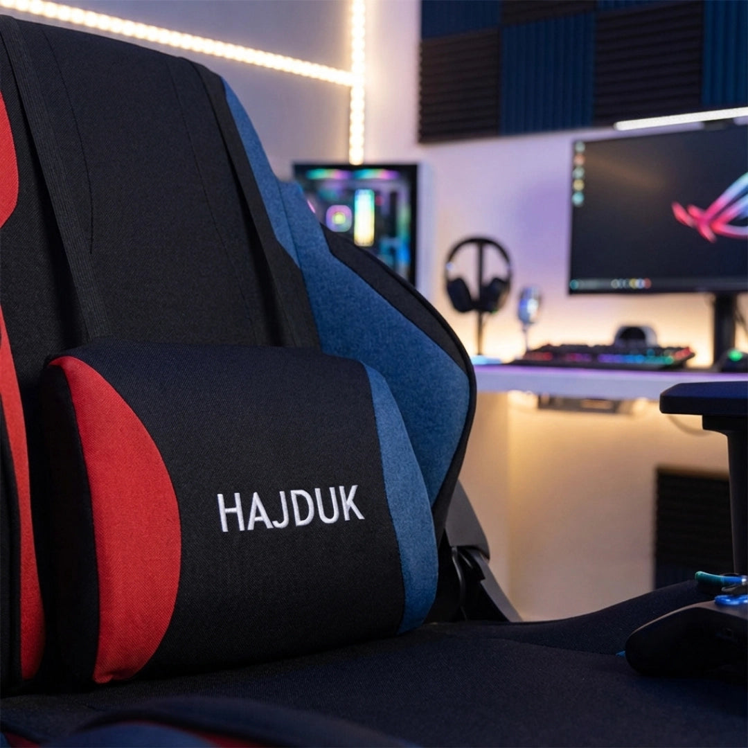 The product is a gaming chair, specifically the UVI X Hajduk model. The image showcases the chair's ergonomic design with a prominent cushion featuring the "HAJDUK" logo, set against a stylish gaming setup that includes a computer monitor and gaming accessories in the background.