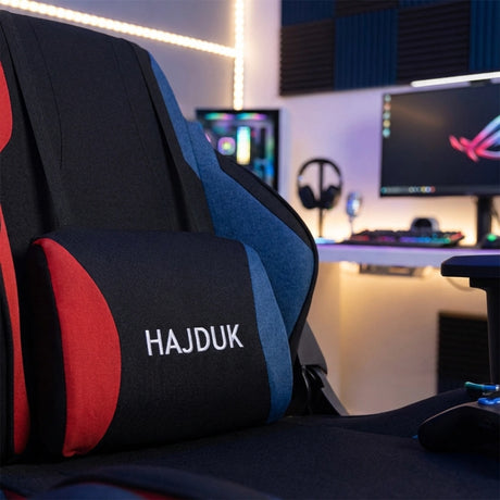 The product is a gaming chair, specifically the UVI X Hajduk model. The image showcases the chair's ergonomic design with a prominent cushion featuring the "HAJDUK" logo, set against a stylish gaming setup that includes a computer monitor and gaming accessories in the background.