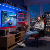 The image features an ergonomic gaming chair, specifically the UVI X Hajduk model. It shows a young woman comfortably seated while playing a racing video game on a large screen. The room is well-lit with ambient lighting, and there are gaming consoles and shelves filled with games in the background.