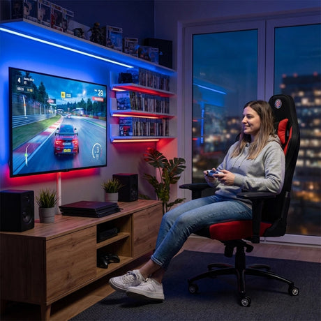 The image features an ergonomic gaming chair, specifically the UVI X Hajduk model. It shows a young woman comfortably seated while playing a racing video game on a large screen. The room is well-lit with ambient lighting, and there are gaming consoles and shelves filled with games in the background.